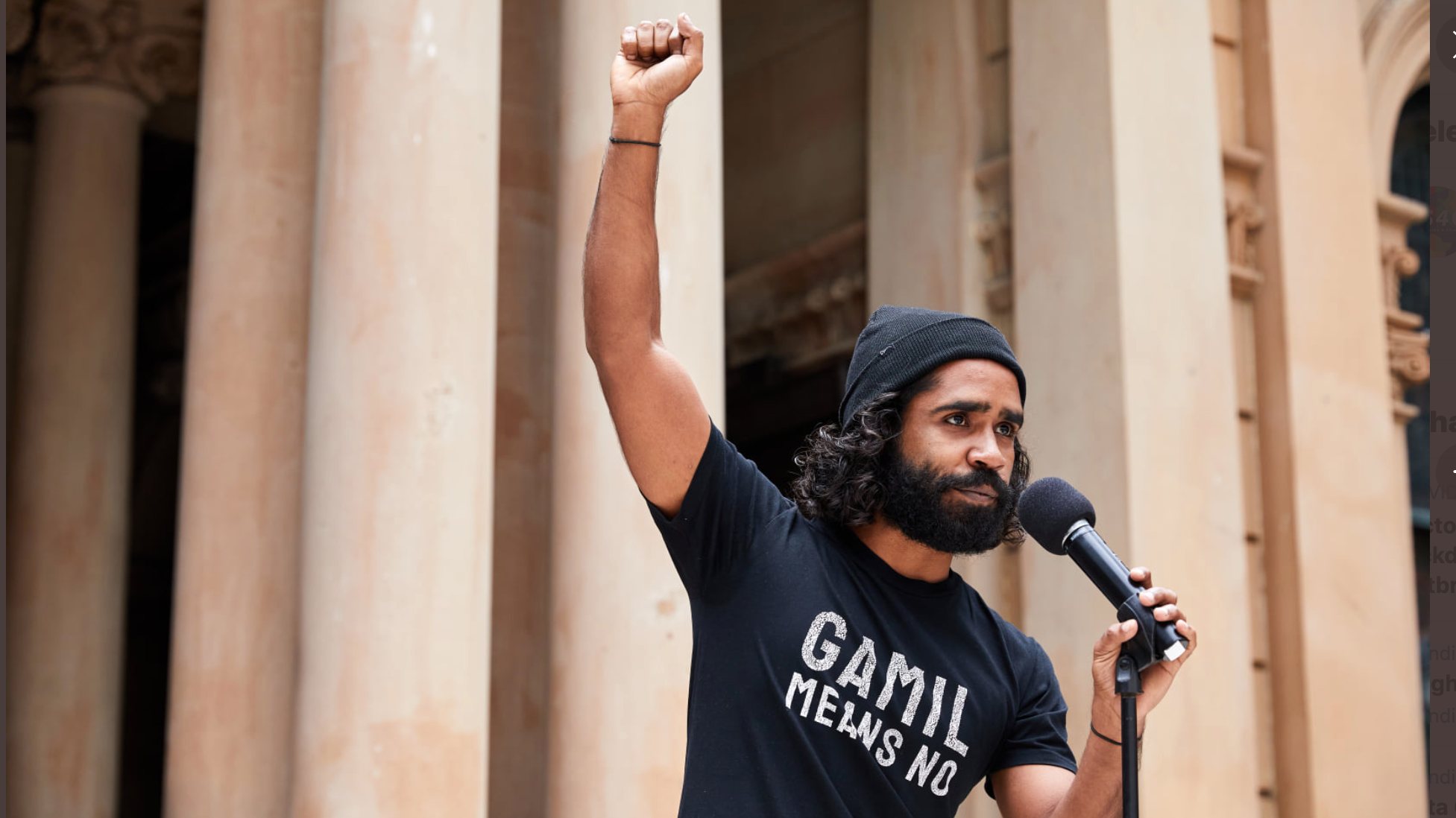Upper torso & headshot of rally speaker Gamilaraay activist Ian Brown wearing a black t-shirt that says “Gamil means No” and holding microphone while raising fist defiantly. His facial features are serious, sad and grim. The background framing (soft focus) is a section of tall concrete columns and archways at the front of the Sydney town hall.