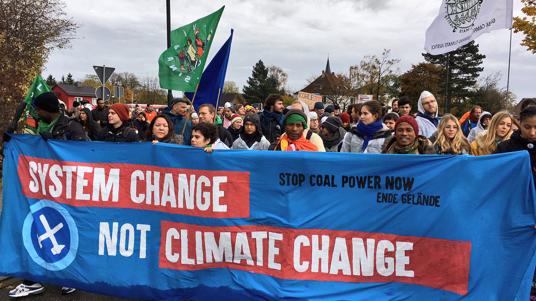 Six POC are carrying a large banner and leading a huge protest march with up to hundred people visible behind them. The banner is blue and red, with black and white text. It says ‘Stop cola power now—Ende Gelände’ and ‘System change, not climate change’, and also depicts the Ende Gelände logo with crossed mallet and pick axe in a circle.