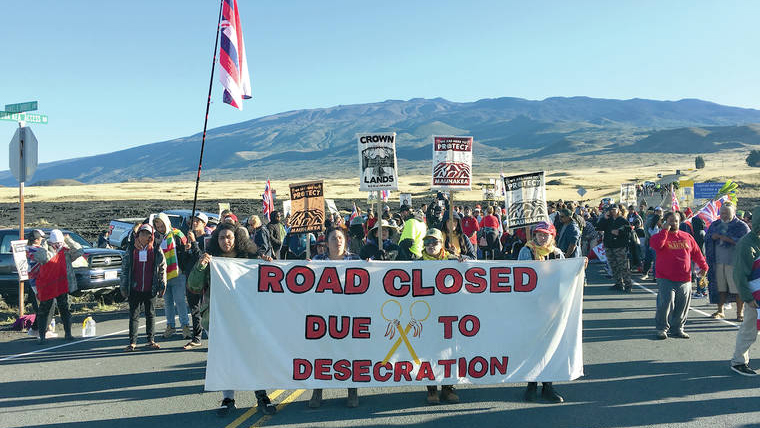 large group of protesters standing in blockade on the orad to Mauna Kea summit holding banners & largest banner reads "Road closed due to desecration"