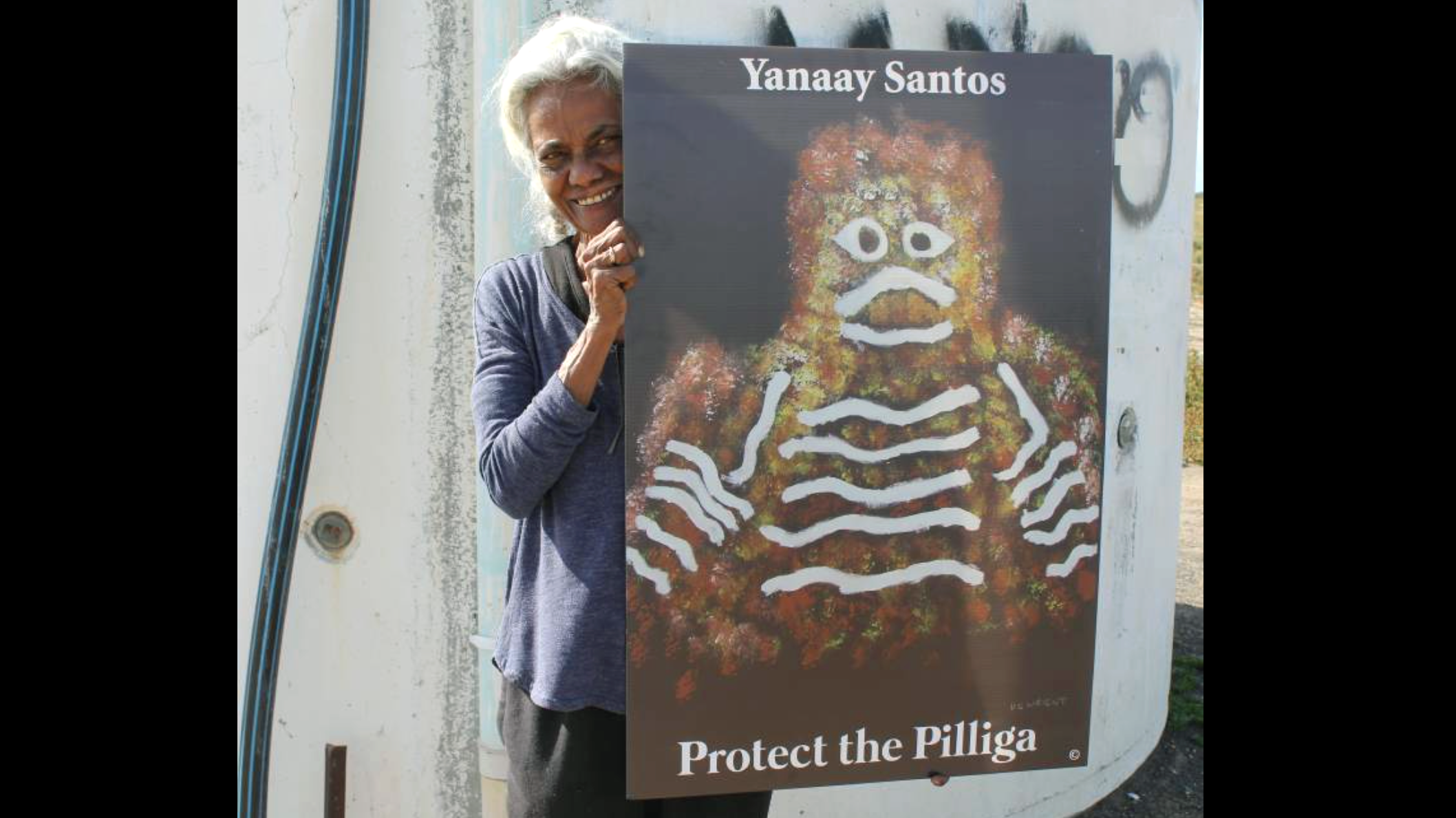 A slender but very strong looking mature Gomeroi Woman is standing in front of a wall that appears to be part of some kind of holding tank. She is holding a sign that says “Yanaay Santos, Protect the Pilliga” and an image invoking a traditional style depiction of the face and shoulders of a humanoid figure using cultural designs. The woman is wearing loose casual clothing (black pants and blue shirt); her hair is silver white and tied back, and she is smiling.