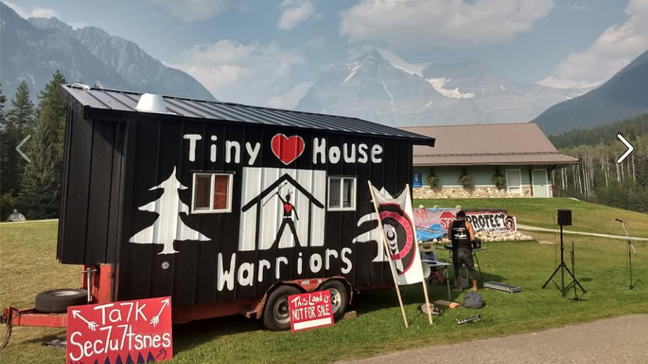 in the foreground is a tiny house on a trailer parked on roadside in a rural area. It has a mural painted on it with Indigenous art symbols and "Tiny House Warriors", striking colours and design. A couple of people are working at a bench beside it with their backs to camera and four protest banners plus other camping equipment is visible. In the background is a wooden building, mountains and blue sky. It is a very idyllic setting.