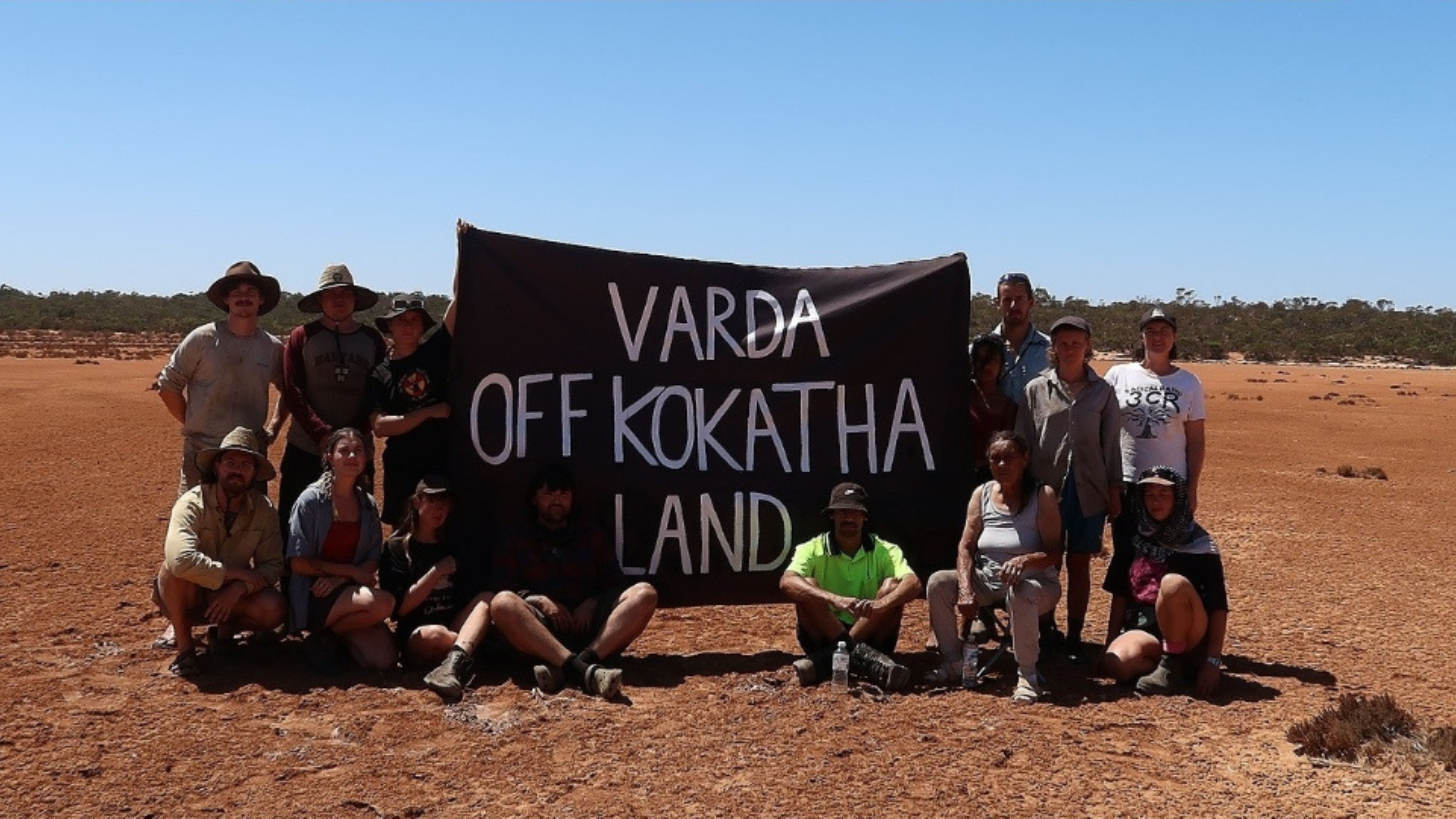 Photo taken on Kokatha Country. In the centre of the photo is a banner which reads "Varda off Kokatha land". Either side of the banner are people involved in the campaigns led by Kokatha Elder Aunty Sue Haseldine, as well as Aunty Sue herself.
