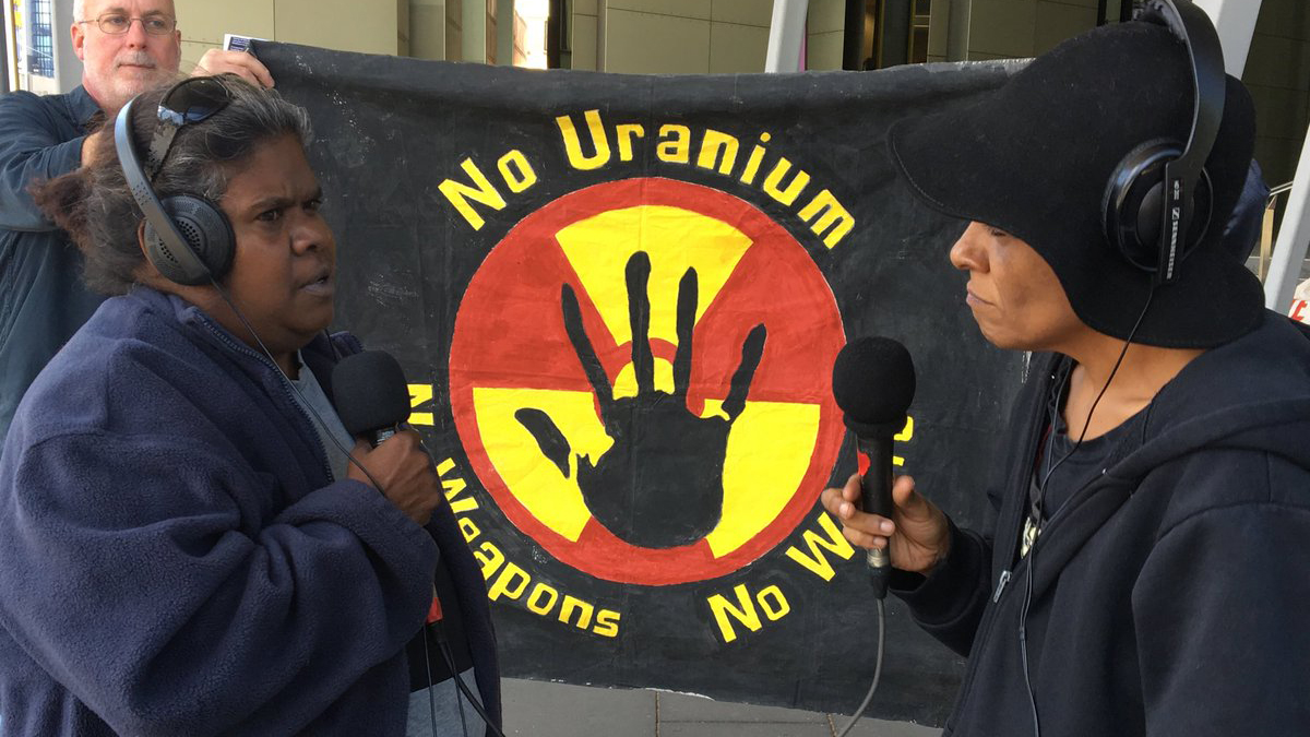 3CR Presenter Viv Malo speaking with Aunty Vicki Abdullah (Tjiwarl Traditional Owner & co-chair of the Australian Nuclear free Alliance at  the IMARC blockade in Melbourne on Oct 28. They are facing each other wearing headphones and holding microphones standing in front of a banner that says "no uranium, no weapons, no way".