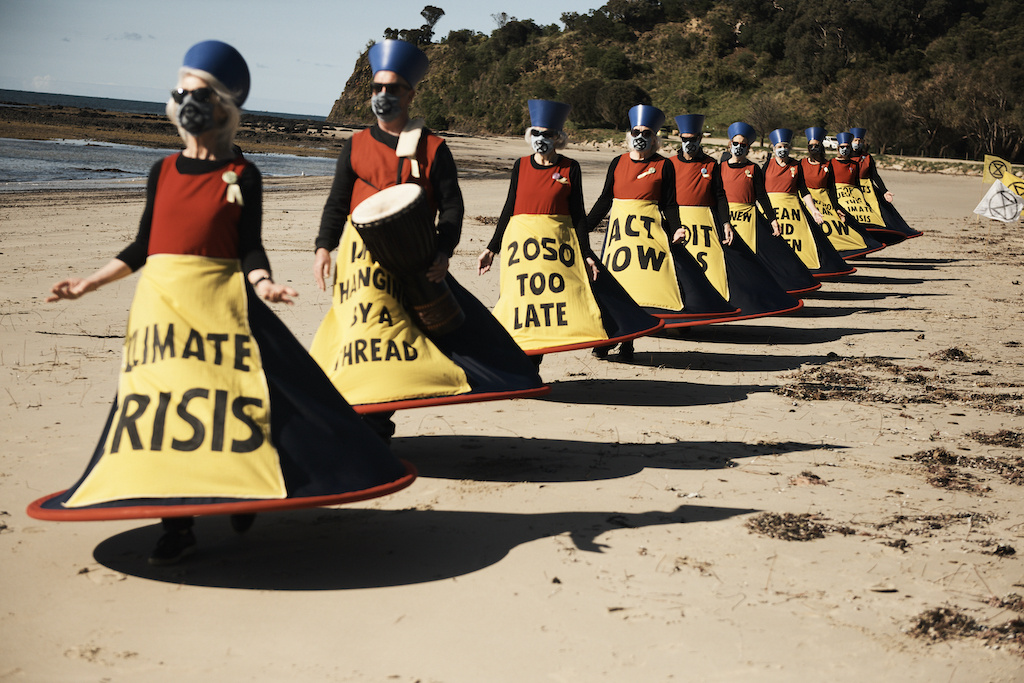 Ten people with blue hats, red tops and blue skirts held out by piping, wearing gas masks. Their yellow aprons carry messages: Climate crisis, Hanging by a thread, 2050 is too late, Act now XR Westernport Sybil Disobedients: Photo provided by Talei Kenyon