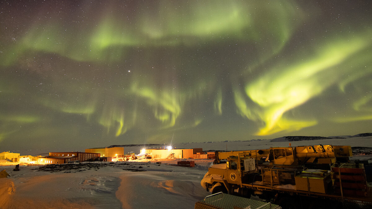 Aurora Australis over Davis station: In the foreground is heavy machinery and buildings and structures of the station with the skyline and playful and intricate light patterns of the aurora bearing down on them 