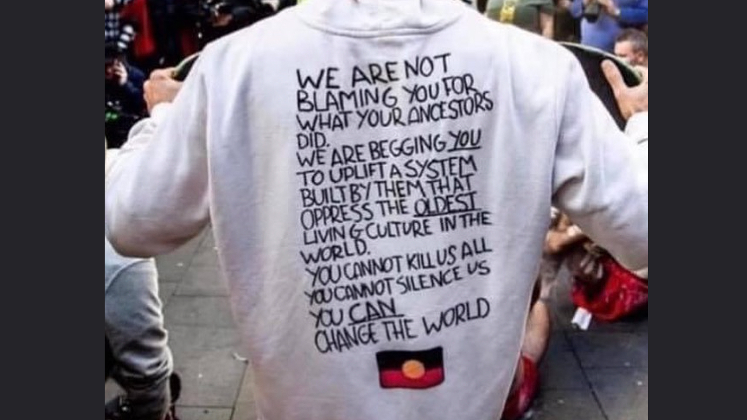 Photograph of protestor's torso from the back. They are wearing a hoodie that says "We are not blaming you for what your ancestors did. We are begging you to uplift a system built by them that oppress the oldest living culture in the world. You can not kill us all. You can not silence us. You can change the world.