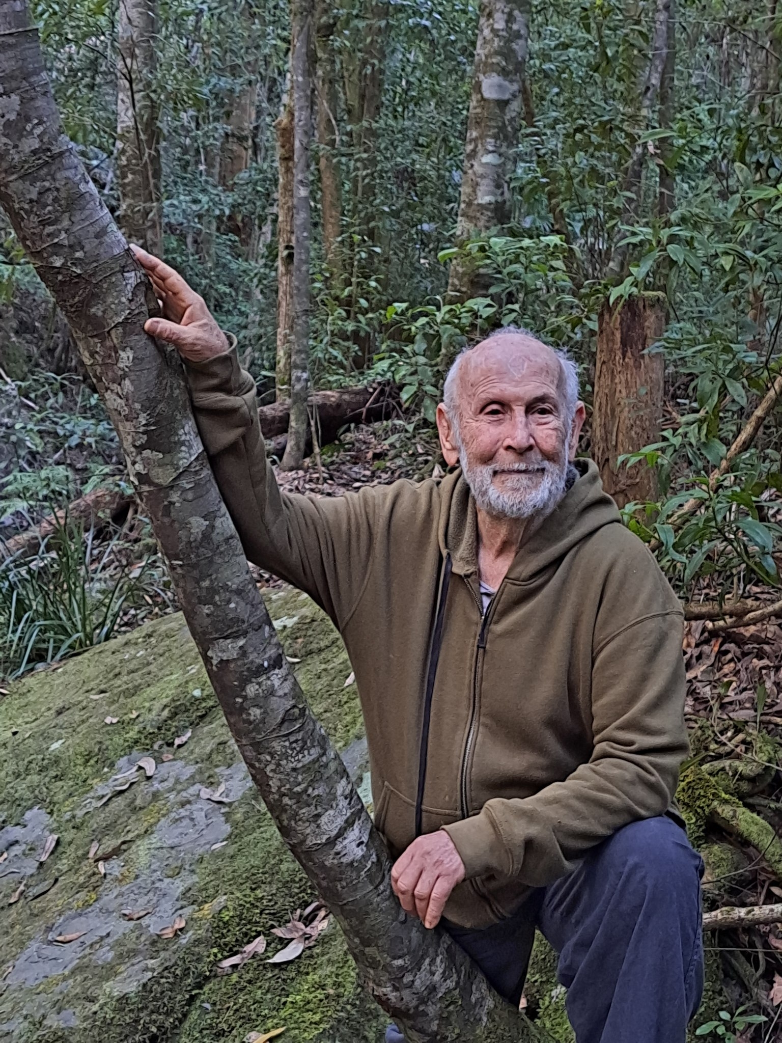 Surrounded by rainforest, John Seed touches a tree with both hands.