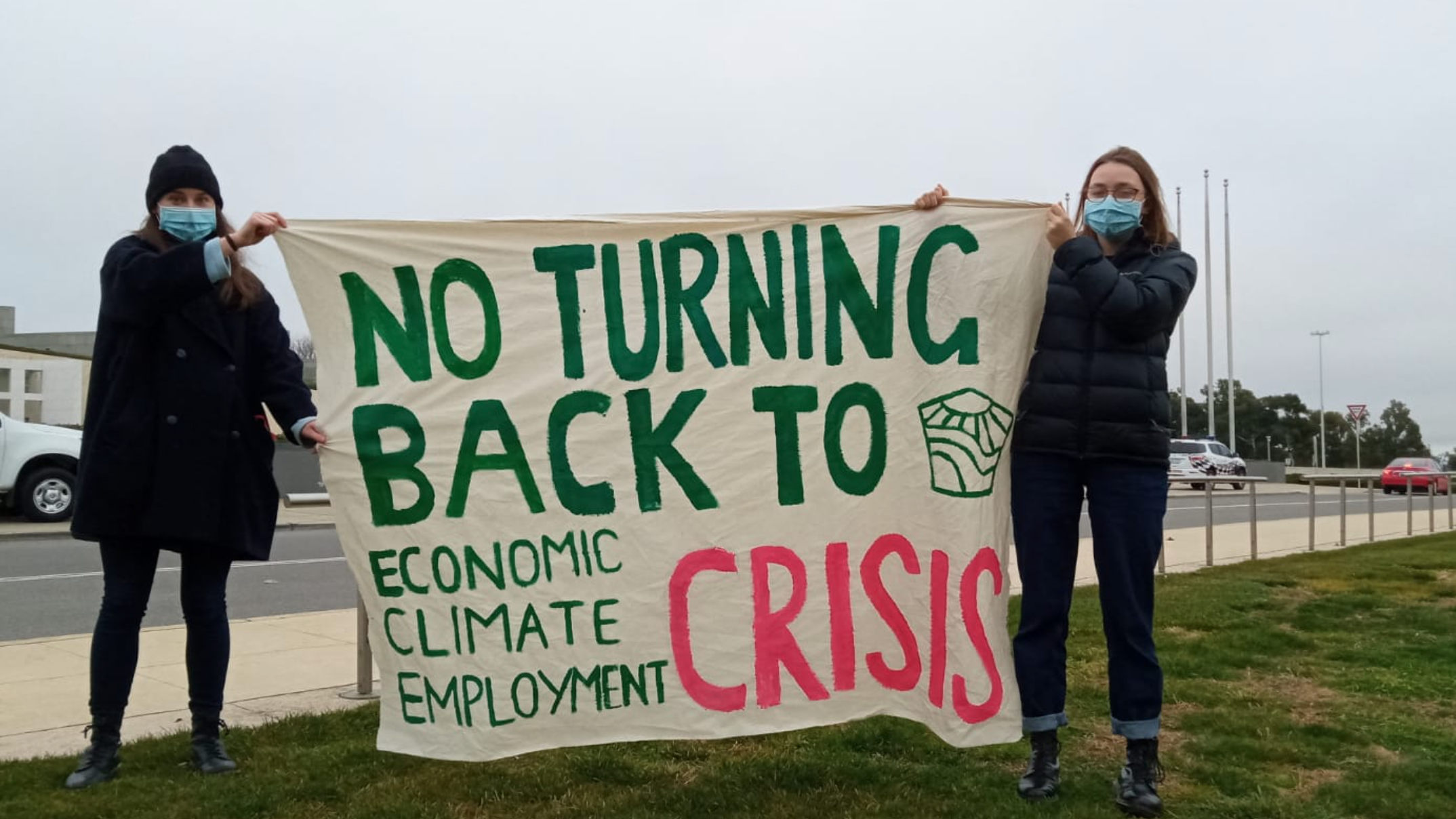 Two young people wearing dark toned warm winter clothes and covid protection face masks are holding a banner up on the side of a busy highway. The banner says “No Turning Back to economic, climate, employment CRISIS”.