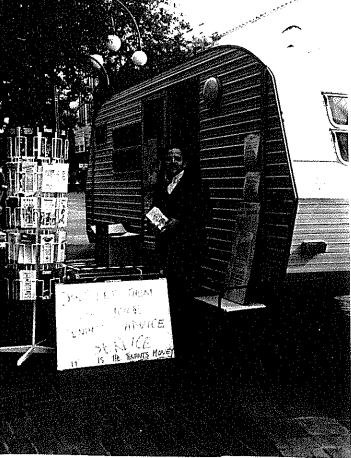 Dark, badly photocopied image showing a man handing out pamphlets outside a caravan in the street.