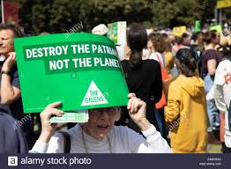 An elderly white woman at a protest holds a sign reading "Destroy the patriarchy, not the planet"