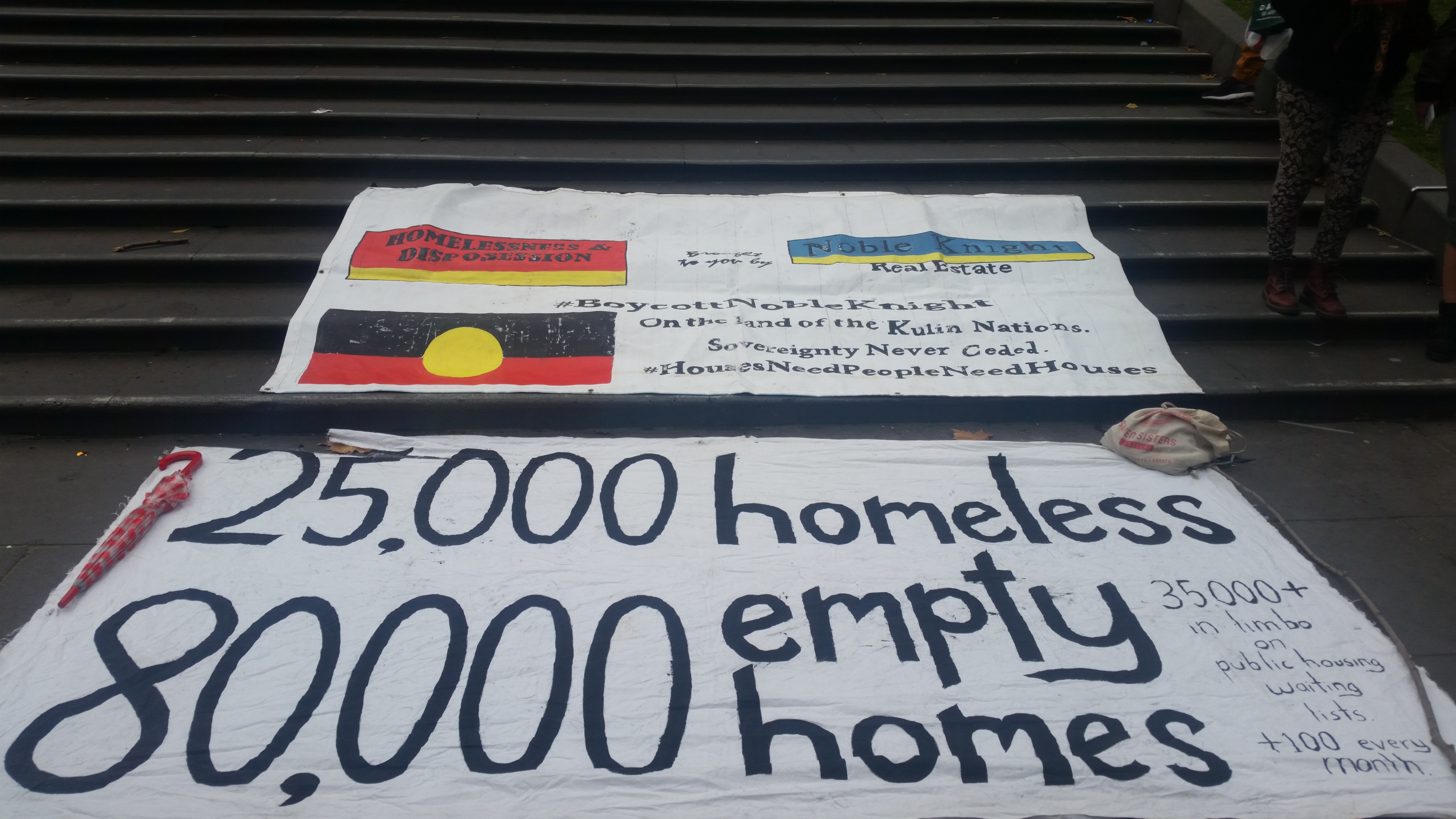 Photo of banners on the state library steps featuring the main one: 25,000 homeless, 80,000 empty homes in big text (in small text 35,000 in limbo on public housing waiting lists +100 every month). There are two other banners in the background, one has the Aboriginal flag and sovereignty never ceded as part of its text. 
