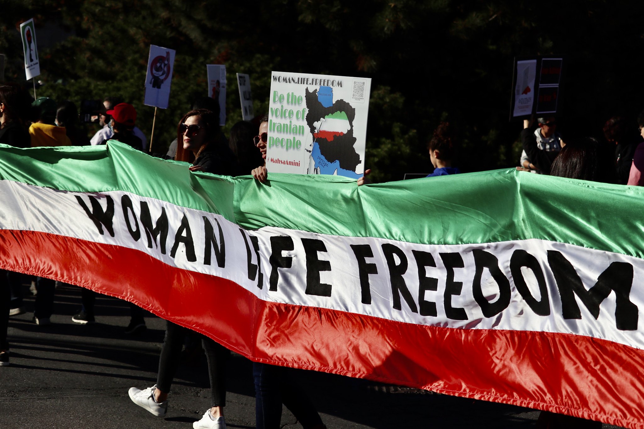 Protesters hold a banner in the colours of the Iranian flag that stretches across the picture with the words 'Women, Life, freedom', in the background people are marching with placards.