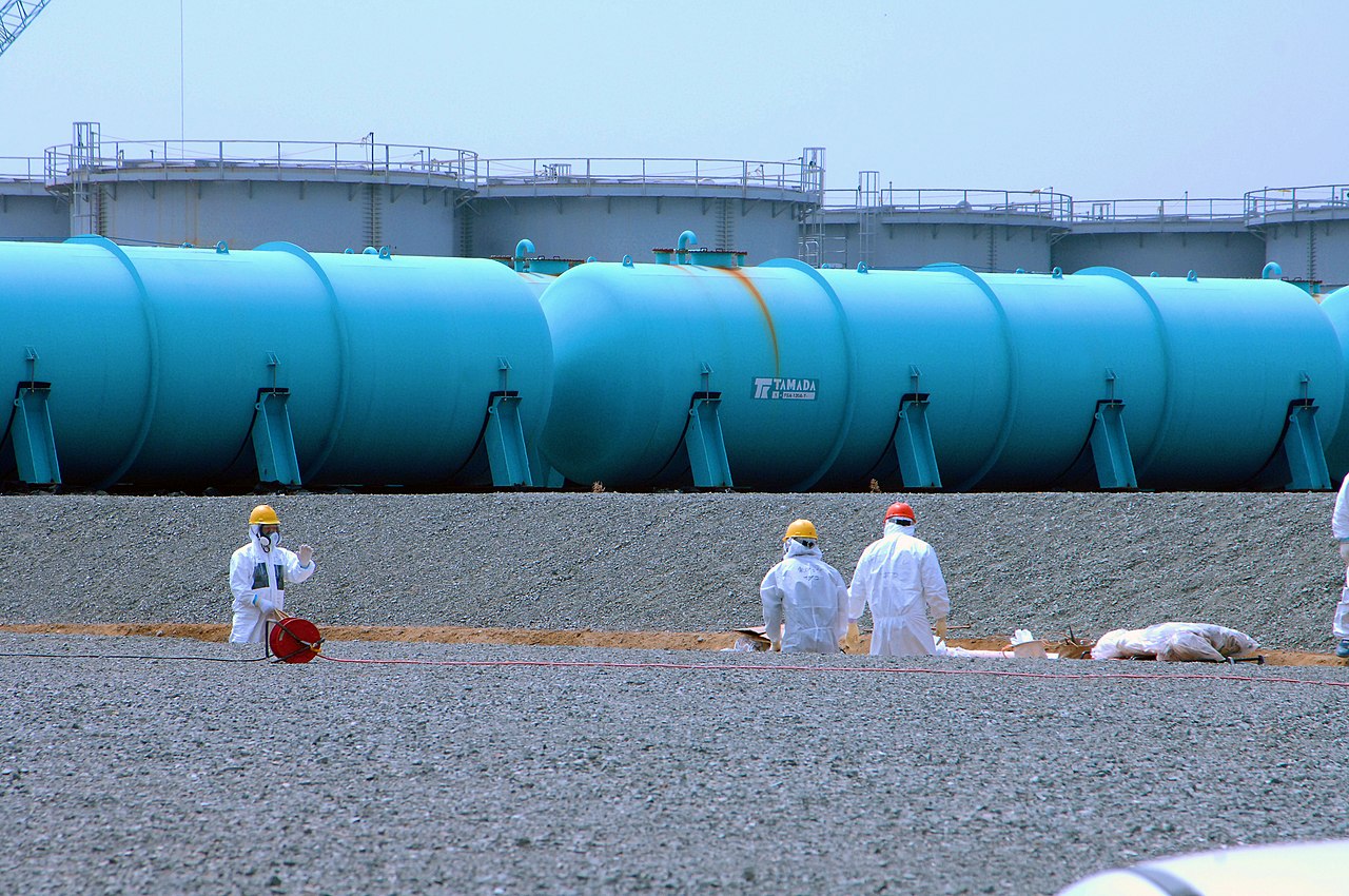 Photo of blue water tanks from Fukushima Daiichi nuclear plant. Tanks are bright blue cylinders and look about 10 metres long and 3 metres high. Three workers in hazmat suits and hard hats stand in gravel in front of the blue tanks. Behind the blue tanks are large upright grey tanks.