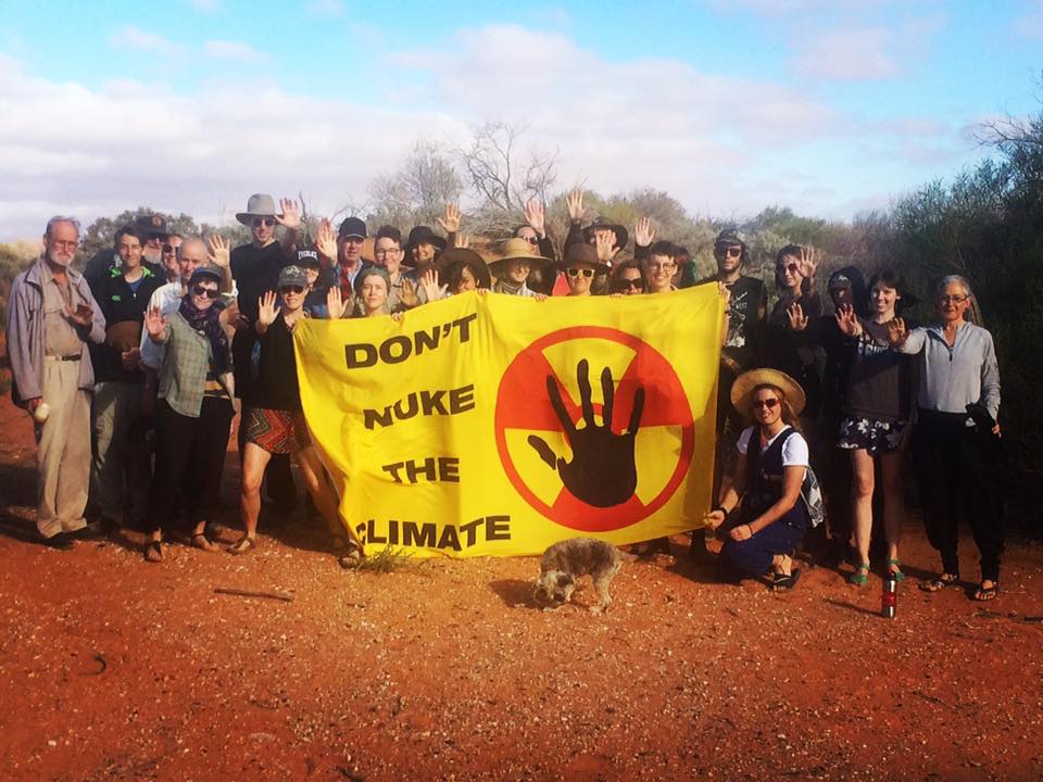 Group of about thirty people holding banner that says "Don't Nuke the Climate", standing in front of sand dunes and bush at Woomera