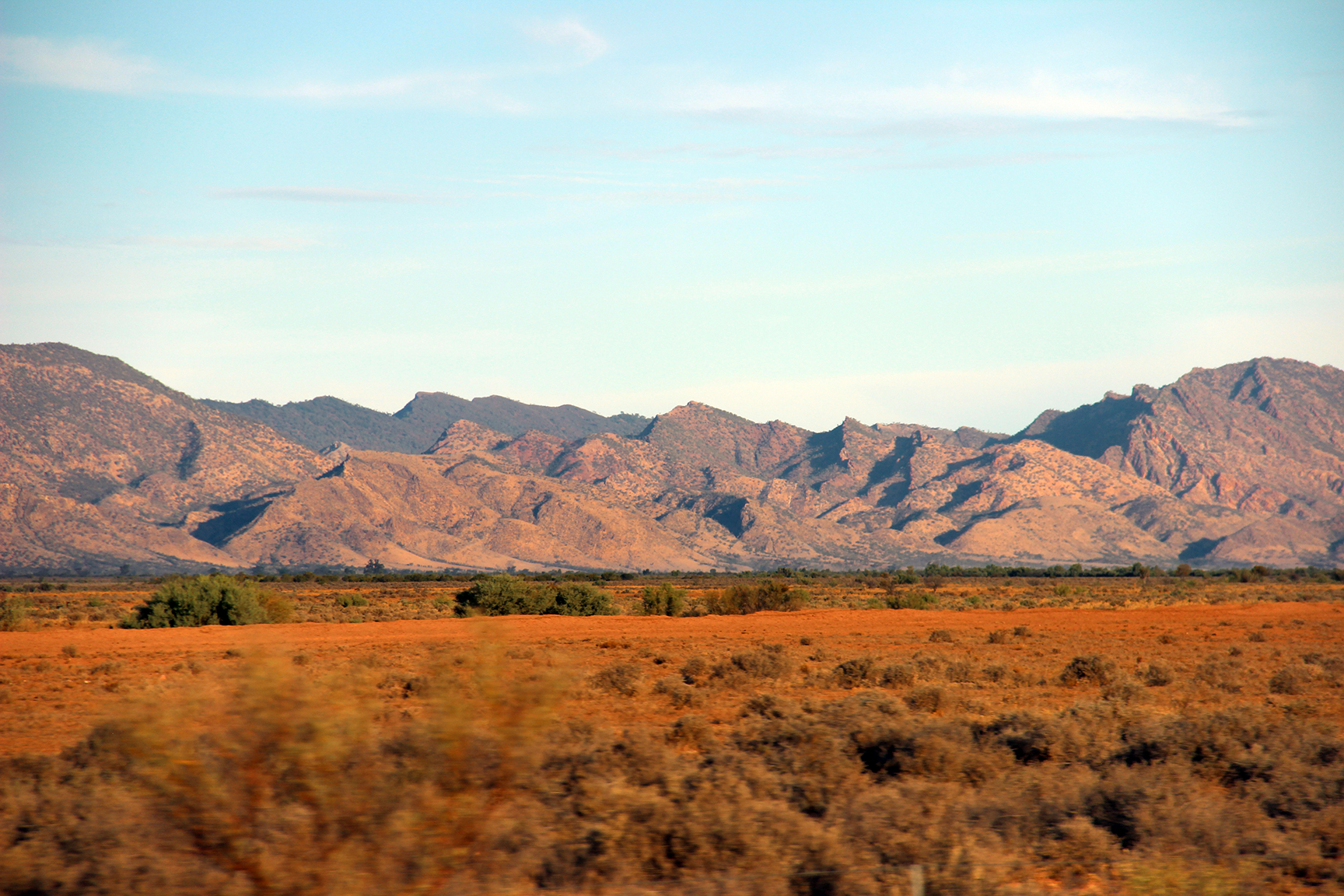 Image taken from moving vehicle, so dry red earth and grasslands in foreground are slightly blurred. Behind are dramatic hills with many peaks, bush covered at the back and less bushy, and dry looking in front.