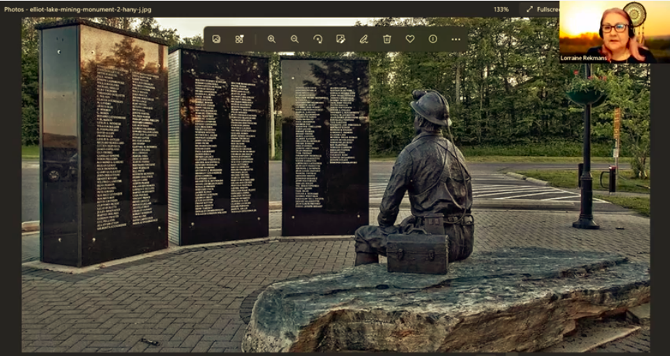 Screenshot from webinar with image of a memorial to miners with three large black square columns at back with names in white on them, and bronze sculpture of miner sitting in foreground. In the top right corner is a small image of Lorraine Rekmans, a middle aged First Nations woman with glasses, pale complexion and pinkish hair.