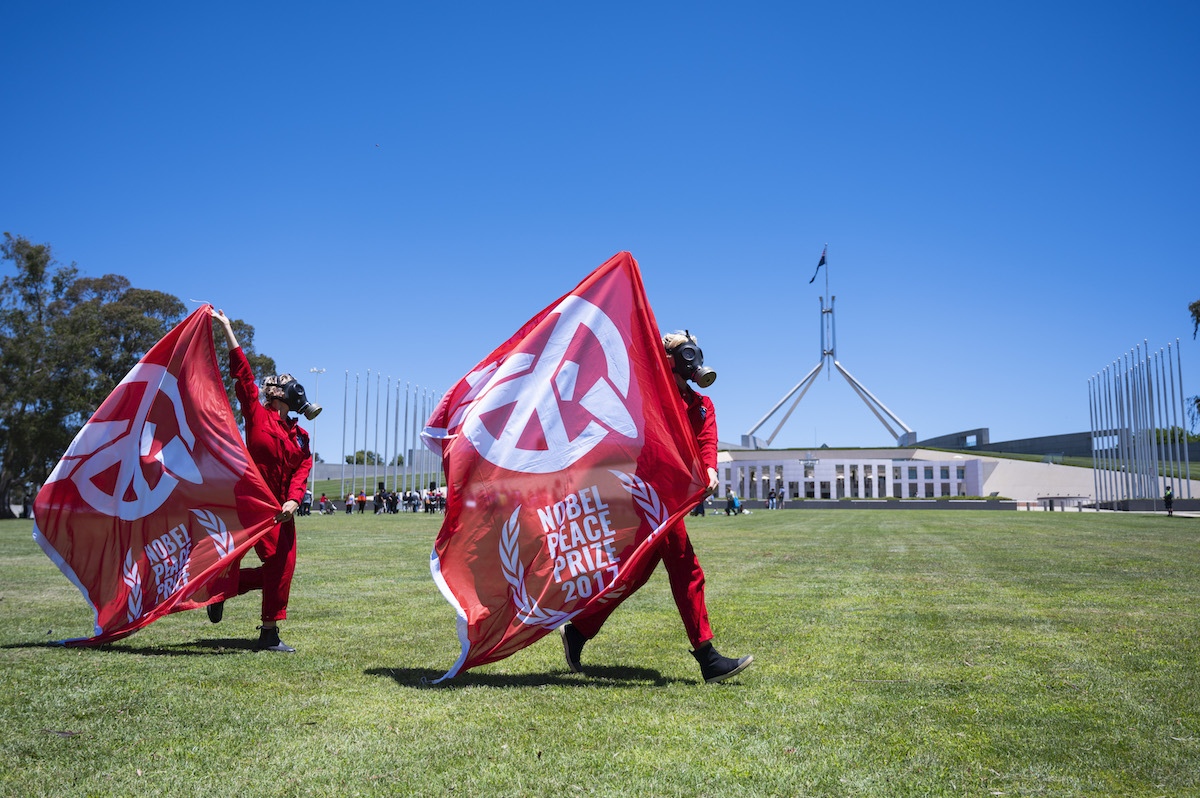 Two people carrying large red ICAN flags walk across lawns in front of the Australian Parliament 