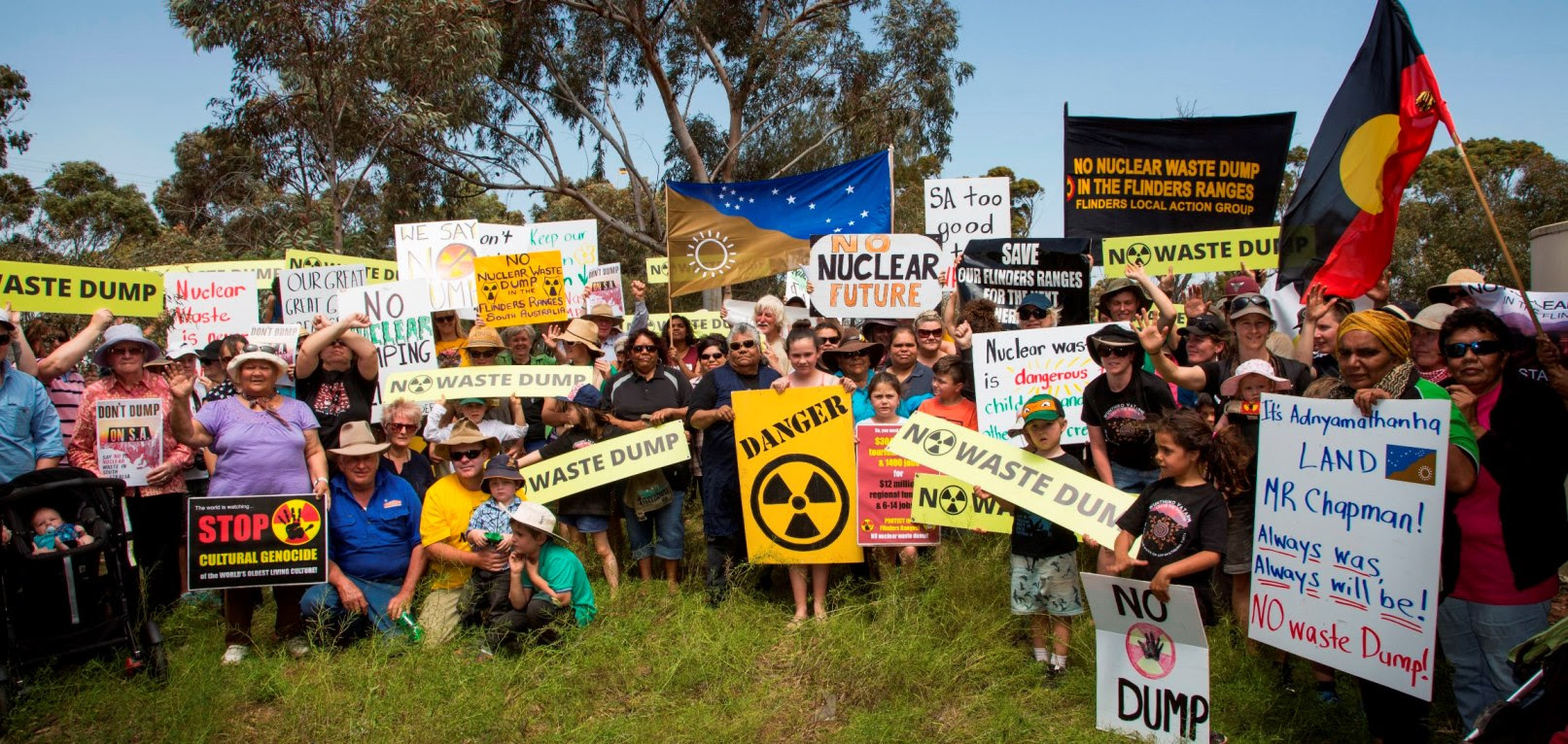 A large crowd of people of all ages outside, in front of a gum tree, holding protest signs that read "No dump", "Danger", "No Cultural genocide", "No nuclear future", "It's Adnyamathanha land Mr Chapman - always was, always will be - No waste dump". There is a Adnyamathanha flag being held up in the centre and an Aboriginal flag held on the right.