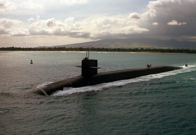Image of large black nuclear submarine in the centre of the photograph, travelling on the surface of sea. There is a forested coast line in the background and a mountain can be seen in the distance as well as a cloudy sky above.