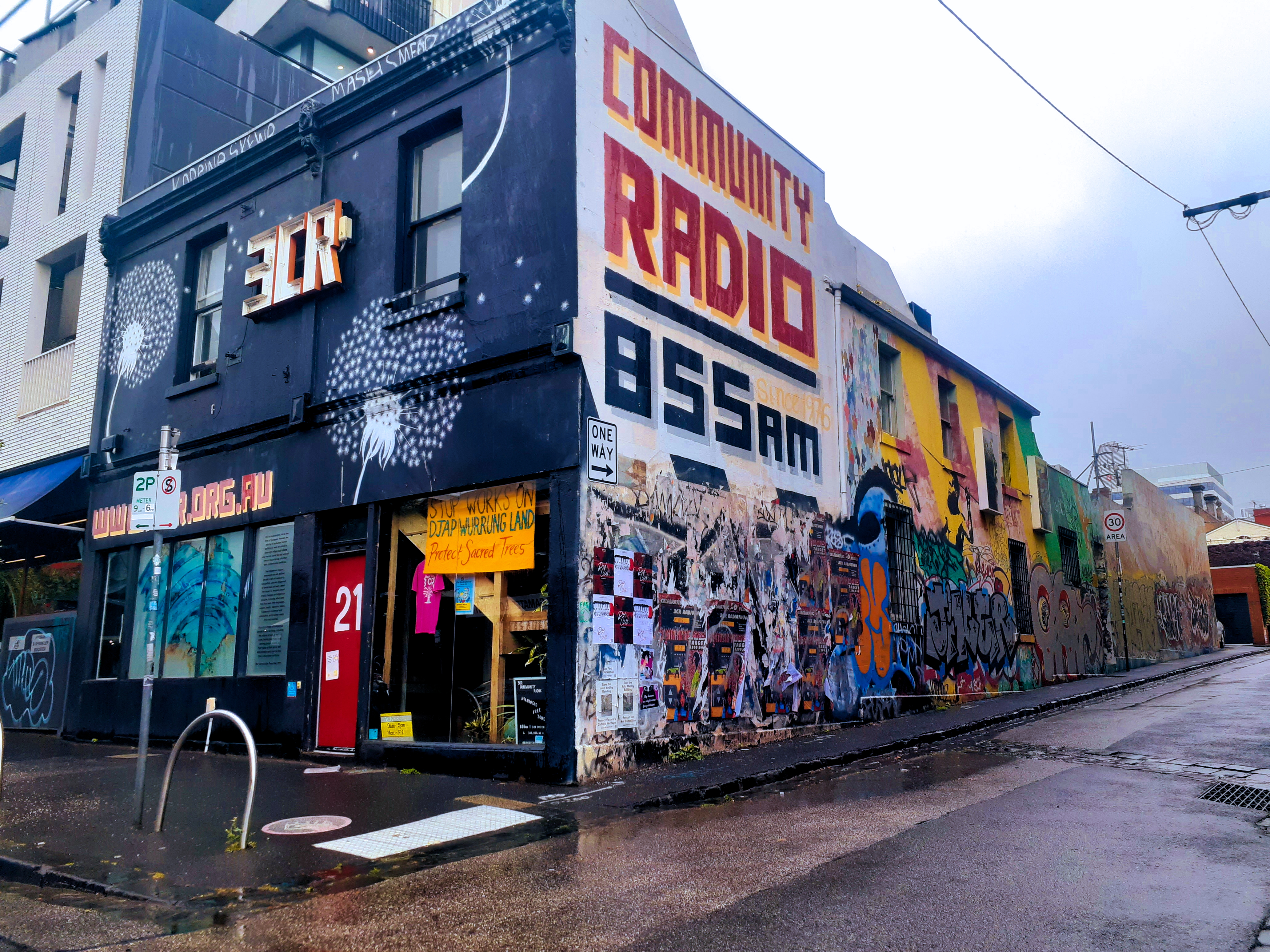 The 3CR building from Smith Street in Fitzroy. The sky is very gray, and the streets wet from rain