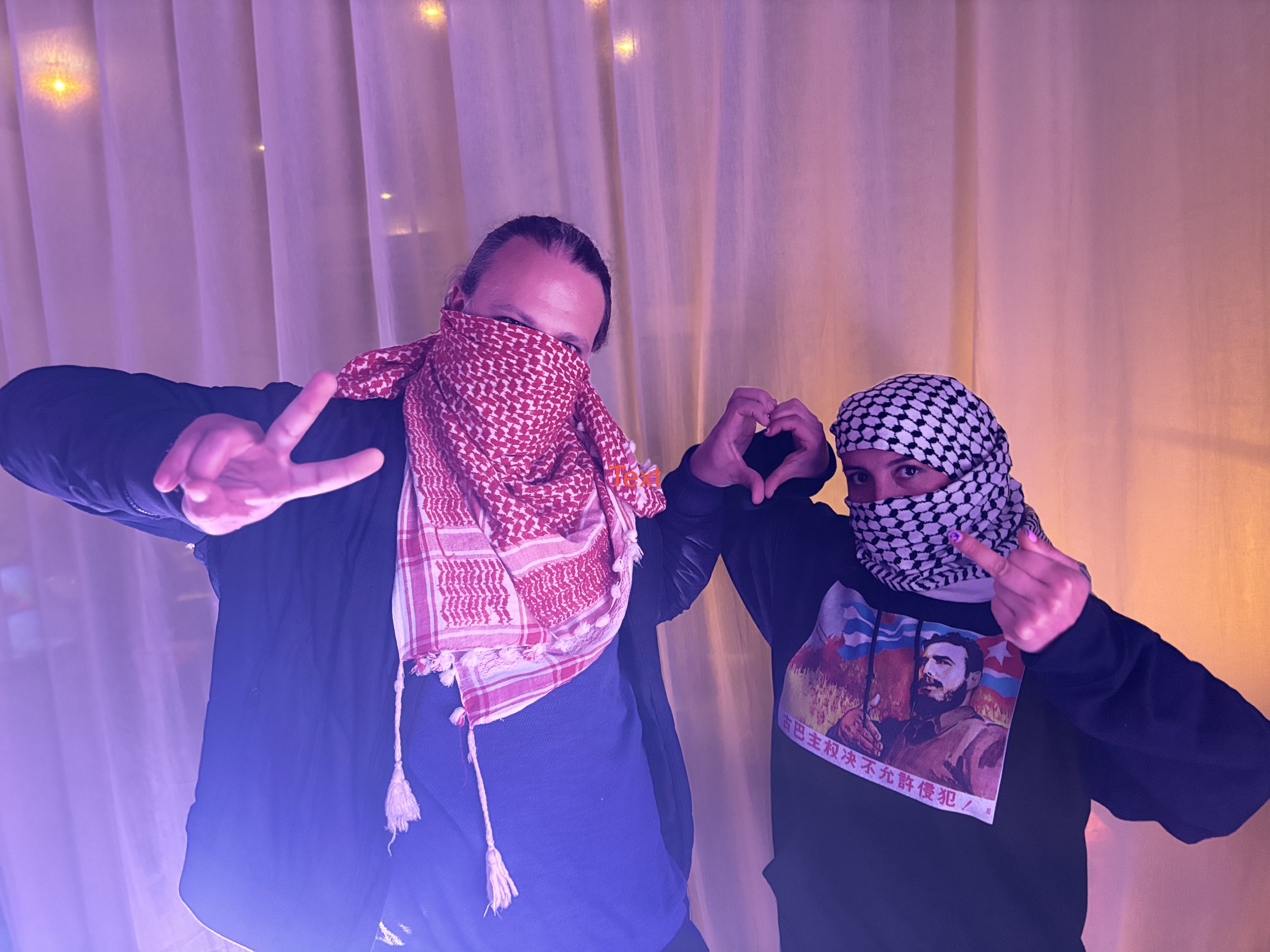 (L-R) Vittorio and Karina wearing Red and Black embroidered Kufiyas, Palestinian Scarves, in front of a semi-transparent curtain, making a love heart out of their respective left and right hands. Vittorio is making a peace symbol with his free hand, while Karina puts up the rude middle finger.