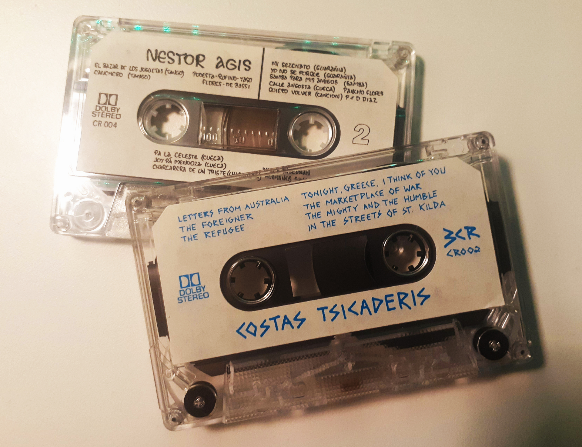 Two cassettes out of their covers. One reads "Costa Tsicaderis", and the other "Nestor Agis", along with track listings and other information.