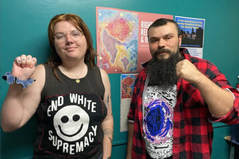 Image description: Em Randall, L, with light-coloured hair, wearing a t shirt that says ‘End White Supremacy’, standing next to Keiran Stewart-Assheton, R. They are in the 3CR studio with fists proudly raised in front of the disability day artwork by Renay Barker-Mulholland.