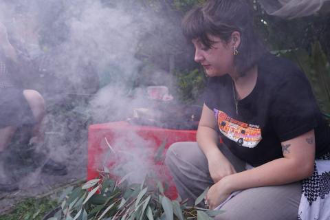 Brittney Henderson squatting next to the sacred flame at a Smoking Ceremony, gazing pensivley into the flame.