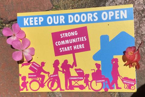 I mage of the post card mentioned in the interview. It features the words 'Keep Our Doors Open, Strong Communities Start Here." Includes image of a diverse looking community entering a neighbourhood house.