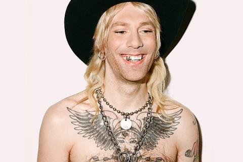 bare-chested person blonde hair black western hat tattoo on chest chains around neck