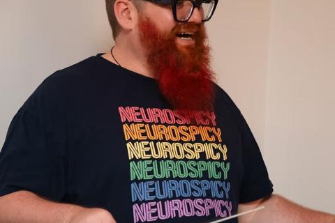 person dark-rimmed glasses beard t-shirt repeating the word neurospicy in rainbow colours