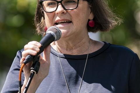 Woman with short dark hair black-rimmed glasses dark top holding microphone