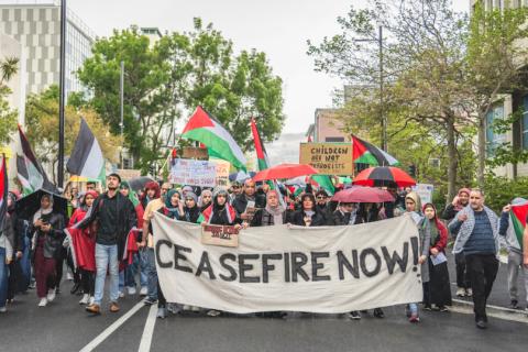 People marching in a pro-Palestine protest with a flag in front that reads 'Ceasfire now'