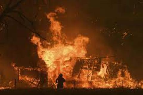 A fire engulfs the structure of a house in a blackened sky. A firefighter stands looking at it in the foreground