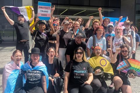 A group of trans rights activists pose and raise their fists and trans flags at a protest outside of the QLD Supreme Court