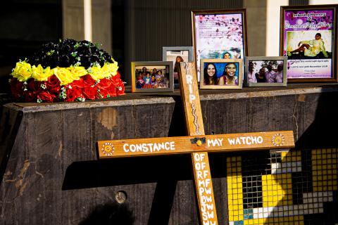 A photograph of the memorial for Constance May Watcho. There is a flower arrangement of black, yellow and red roses, a cross with Ms Watcho's name and designs painted on it, and a cluster of framed photographs of Ms Watcho and her family.