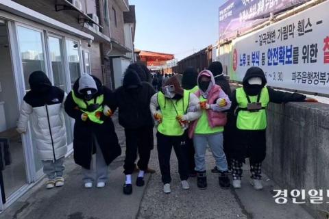 Image of five protesters locking arms in Paju's red light district in South Korea. They are wearing yellow neon vests and covering their face.