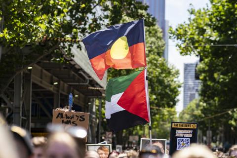 A photograph of a flag pole flying an Aboriginal flag above a Palestinian flag, being held by someone standing in the crowd during Melbourne's 2023 Invasion Day rally.
