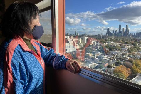 A Vietnamese resident of the housing estate looks out the window of her flat. You can see a view of Narrm. 