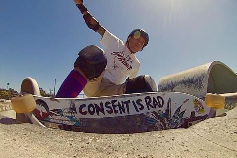 A low-angle photograph of a skateboarder in the middle of a trick on the rim of a skate park bowl. You can see the bottom of their board, which has a large sticker on it reading "Consent is Rad".