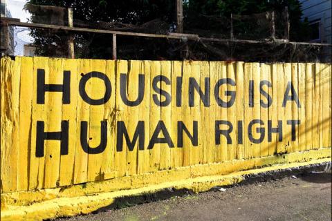 A high wooden fence painted bright yellow, with the words 'housing is a human right' painted over the top in large black capital letters.