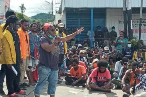 A photograph of West Papuan feminist Esther Haluk speaking at a peaceful demonstration against the break up of the provinces. She is standing to the left of the photograph, speaking into a microphone and gesturing to a crowd seated on the ground.