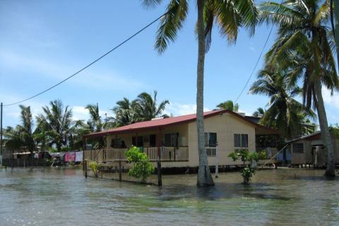 A photograph of a king tide on Saibai Island from 2010. A building on stilts is surrounded by high seawater, covering the ground and almost reaching the base of the building. Fences and plants in the foreground are partially submerged, and other buildings on stilts are visible in the background.