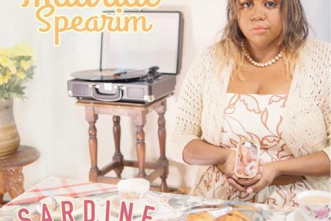 The cover for Maurial Spearim's single Sardine Baby. Maurial, dressed in 1950s femme style, sits at a table holding a Sardine Baby (tin with a pebble and cloth). The table is laid for tea, with a plate of scones, teacups, and more Sardine Babies. In the background is a record player on a small table.