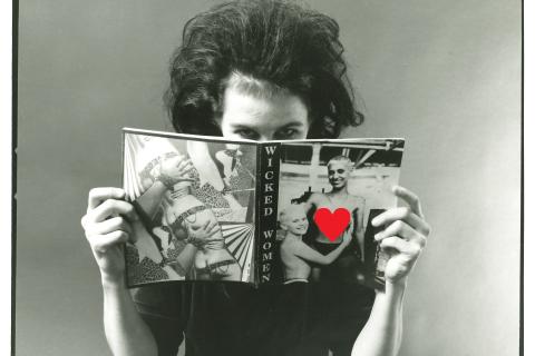 Black and white portrait style image showing a woman with teased hair and black eyeliner in a black velvet dress reading a copy of Wicked Women magazine. She holds the magazine in front of her face with just her eyes looking out at us, disrupting the relationship between viewer and viewed