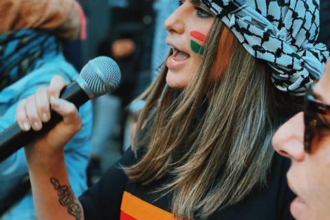 Palestinian activist, campaigner, and storyteller Jeanine Hourani speaks to a microphone at a protest. She is wearing a black and white kuffiyeh (كُوفِيَّة‎) with a black, yellow and red Always Was (Aboriginal land) sweater