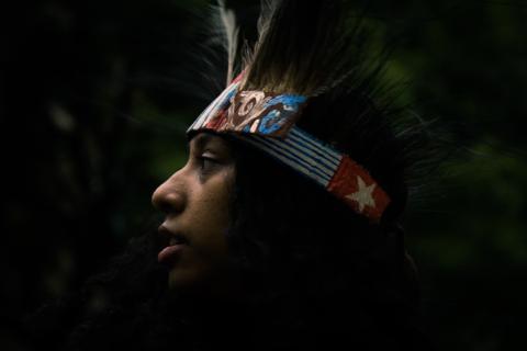 Image of a young West Papuan woman looking out to the distance wearing a West Papuan headdress with feathers and featuring the West Papuan morning star flag.