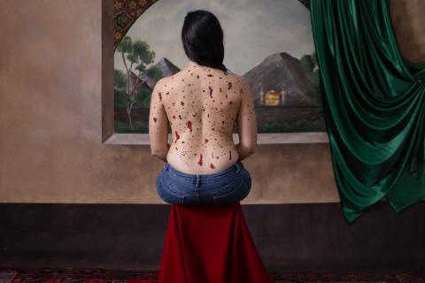 image of a painted background inside a house. There is a woman sitting on on a red stool looking out the window. Her back is faced towards us and is covered in red jewels depicting blood.