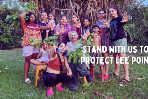 A group of Larrakia women and children pose, painted up, in front of lush greenery. Overlaid on the photo is the text "stand with us to save Lee Point."