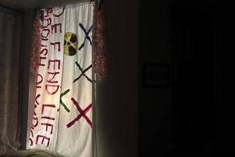An anti-nuclear banner reading DEFEND LIFE hangs illuminated in a window against a dark background